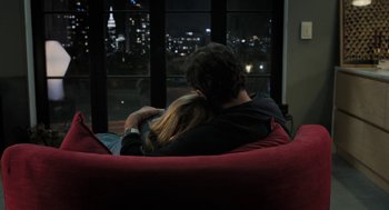 Movie still from “Holy Lands” (2017), directed by Amanda Sthers – A man and a woman sitting on a red couch looking out of a window at night; Medium shot, High angle