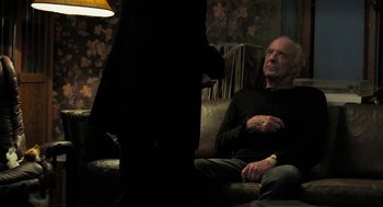 Movie still from “Holy Lands” (2017), directed by Amanda Sthers – An older man sitting on a couch talking to a woman; Medium shot, Over the shoulder angle