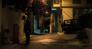 Movie still from “Holy Lands” (2017), directed by Amanda Sthers – A man in a suit and hat standing on the side of the street; Wide shot, High angle