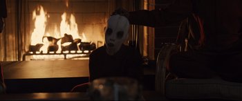 Movie still from “Us” (2019), directed by Jordan Peele – A person wearing a mask sitting in front of a fire place; Medium shot, Over the shoulder angle