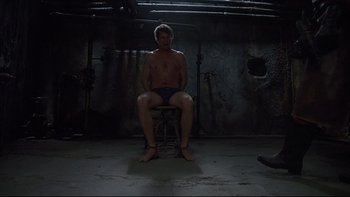 Movie still from “Hostel” (2005), directed by Eli Roth – A man sitting on a chair in a dark room; Wide shot, High angle