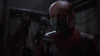 Movie still from “Hostel” (2005), directed by Eli Roth – A man wearing a surgical face mask in a dark room; Medium shot, Low angle