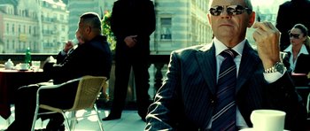 Movie still from “Hostel: Part II” (2007), directed by Eli Roth – A man in a suit and tie sitting on a chair; Medium shot, Low angle