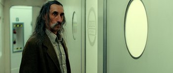 Movie still from “Hostel: Part II” (2007), directed by Eli Roth – A man with long grey hair and a long beard standing in front of a door; Medium shot, Over the shoulder angle