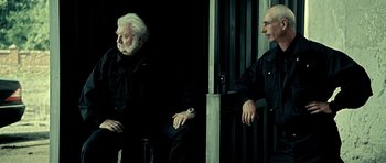 Movie still from “Hostel: Part II” (2007), directed by Eli Roth – Two men sitting next to each other in front of a building; Medium shot, Low angle