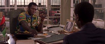 Movie still from “Hotel Rwanda” (2004), directed by Terry George – A man sitting at a table holding a cigar in his hand; Medium shot, Over the shoulder angle