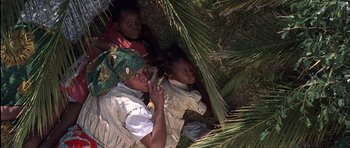 Movie still from “Hotel Rwanda” (2004), directed by Terry George – A group of people laying in the grass next to a palm tree; Medium shot, High angle