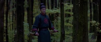 Movie still from “House of Flying Daggers” (2004), directed by Yimou Zhang – A man holding two wooden swords in a forest; Medium shot, Over the shoulder angle