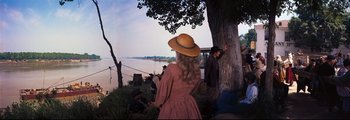 Movie still from “How the West Was Won” (1962), directed by George Marshall – A woman wearing a dress and a hat standing next to a tree near a body of water; Wide shot, Over the shoulder angle