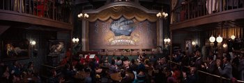 Movie still from “How the West Was Won” (1962), directed by George Marshall – People are sitting at tables in front of a stage with an image on the wall; Extreme Wide shot, High angle