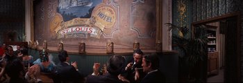 Movie still from “How the West Was Won” (1962), directed by George Marshall – A group of men standing around each other in front of a painting; Wide shot, Over the shoulder angle
