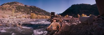 Movie still from “How the West Was Won” (1962), directed by George Marshall – A man in a hat is sitting on a rock by a river; Wide shot, Low angle