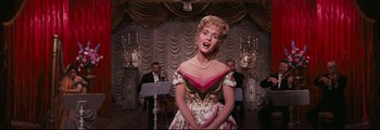 Movie still from “How the West Was Won” (1962), directed by George Marshall – A woman wearing a dress and a pearl necklace in front of a microphone; Medium shot, Low angle