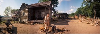 Movie still from “How the West Was Won” (1962), directed by George Marshall – A man standing in front of a log cabin with two dogs; Wide shot, Low angle