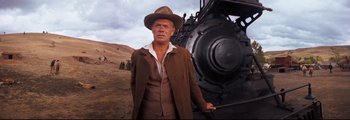 Movie still from “How the West Was Won” (1962), directed by George Marshall – A man standing next to an old fashioned train; Medium shot, Low angle