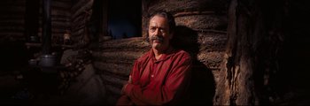 Movie still from “How the West Was Won” (1962), directed by George Marshall – A man in a red shirt sitting in front of a log wall; Medium shot, Low angle