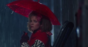 Movie still from “Howard the Duck” (1986), directed by Willard Huyck – A woman holding an umbrella while sitting in a chair; Medium shot, Low angle