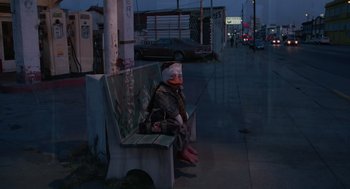 Movie still from “Howard the Duck” (1986), directed by Willard Huyck – A person sitting on a bench on the side of the street; Wide shot, High angle