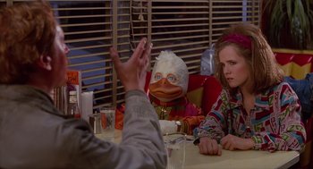 Movie still from “Howard the Duck” (1986), directed by Willard Huyck – A person sitting at a table talking to a person sitting next to a duck puppet; Medium shot, Over the shoulder angle