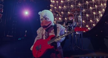Movie still from “Howard the Duck” (1986), directed by Willard Huyck – A person in a chicken mask holding a guitar; Medium shot, Low angle