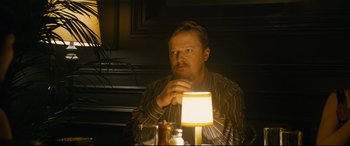Movie still from “Hustlers” (2019), directed by Lorene Scafaria – A man sitting in front of a lamp in a dimly - lit room; Medium shot, Over the shoulder angle