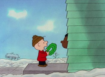 Movie still from “It's Christmastime Again, Charlie Brown” (1992), directed by Bill Melendez – A charlie brown character holding a wreath in front of a building; Extreme Wide shot, High angle