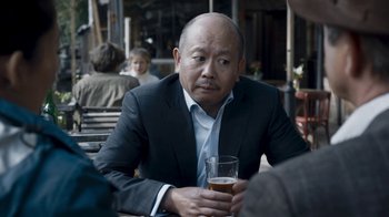 Movie still from “Killing Eve” (2018), created by Phoebe Waller-Bridge – A man sitting at a table holding a glass of beer; Close Up shot, Over the shoulder angle