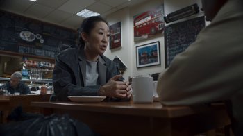 Movie still from “Killing Eve” (2018), created by Phoebe Waller-Bridge – A woman sitting at a table with a cup of coffee; Medium shot, Over the shoulder angle
