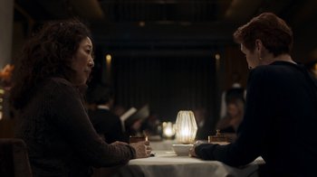 Movie still from “Killing Eve” (2018), created by Phoebe Waller-Bridge – Two people are sitting at a table in front of a lamp; Medium shot, Over the shoulder angle