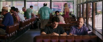 Movie still from “War Dogs” (2016), directed by Todd Phillips – A man sitting at a table in front of other people; Medium shot, Over the shoulder angle