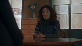 Movie still from “Killing Eve” (2018), created by Phoebe Waller-Bridge – A woman sitting at a table in front of a fan; Medium shot, Over the shoulder angle