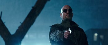 Movie still from “War for the Planet of the Apes” (2017), directed by Matt Reeves – A bald man wearing sunglasses holding a gun in his hand; Close Up shot, Low angle