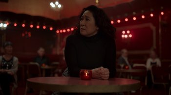 Movie still from “Killing Eve” (2018), created by Phoebe Waller-Bridge – A woman sitting in front of a candle in a dark room; Medium shot, Over the shoulder angle