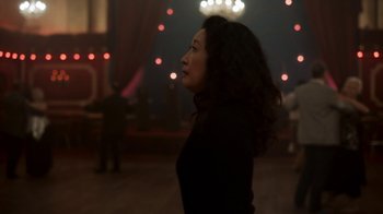 Movie still from “Killing Eve” (2018), created by Phoebe Waller-Bridge – A woman standing in a room with lights in the background; Medium shot, Over the shoulder angle