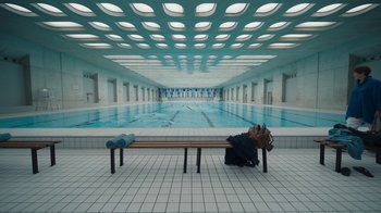 Movie still from “Killing Eve” (2018), created by Phoebe Waller-Bridge – A person sitting on top of a bench in front of an indoor swimming pool; Extreme Wide shot, High angle