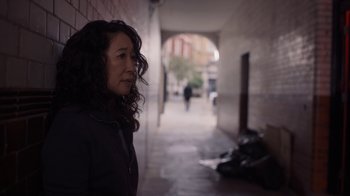Movie still from “Killing Eve” (2018), created by Phoebe Waller-Bridge – A woman standing on a sidewalk near a brick wall; Close Up shot, Over the shoulder angle