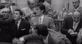 Movie still from “L'Eclisse” (1962), directed by Michelangelo Antonioni – Black and white photograph of a man smoking a cigarette in front of other men; Medium shot, Over the shoulder angle