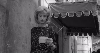 Movie still from “L'Eclisse” (1962), directed by Michelangelo Antonioni – A woman standing in front of a building holding a piece of paper; Medium shot, Low angle