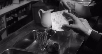 Movie still from “L'Eclisse” (1962), directed by Michelangelo Antonioni – A person holding a paper with a drawing on it next to a glass of water; Close Up shot, Over the shoulder angle