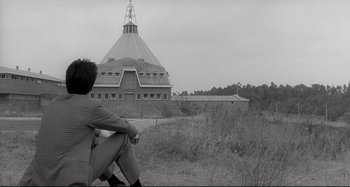 Movie still from “L'Eclisse” (1962), directed by Michelangelo Antonioni – A man sitting on the ground in front of a building; Wide shot, Low angle