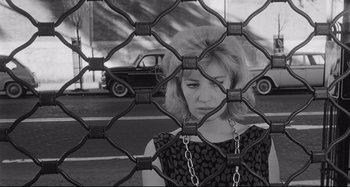 Movie still from “L'Eclisse” (1962), directed by Michelangelo Antonioni – A woman standing in front of a metal fence with cars in the background; Medium shot, Low angle