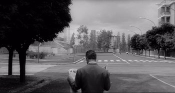 Movie still from “L'Eclisse” (1962), directed by Michelangelo Antonioni – A man reading a newspaper while standing on the side of the road; Extreme Wide shot, High angle