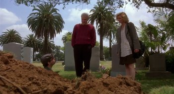 Movie still from “L.A. Story” (1991), directed by Mick Jackson – Two people and a man standing in front of a pile of dirt in a cemetery; Wide shot, Over the shoulder angle