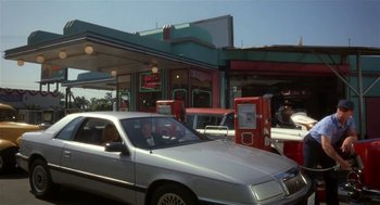 Movie still from “L.A. Story” (1991), directed by Mick Jackson – A man sitting in the drivers seat of a car at a gas station; Wide shot, Low angle