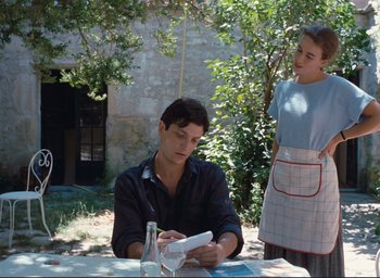 Movie still from “La Belle Noiseuse” (1991), directed by Jacques Rivette – A man sitting at a table with a woman standing next to him; Medium shot, Over the shoulder angle