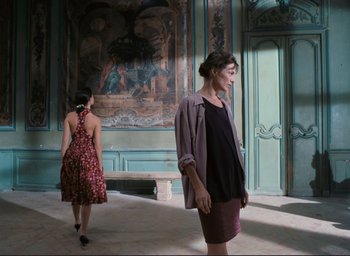 Movie still from “La Belle Noiseuse” (1991), directed by Jacques Rivette – Two women are standing in a room in front of a mural; Wide shot, Over the shoulder angle