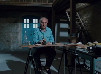 Movie still from “La Belle Noiseuse” (1991), directed by Jacques Rivette – An older man sitting at a table in front of an open door; Wide shot, Low angle