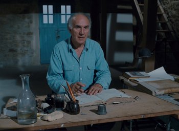Movie still from “La Belle Noiseuse” (1991), directed by Jacques Rivette – An older man sitting at a table in front of papers; Medium shot, High angle