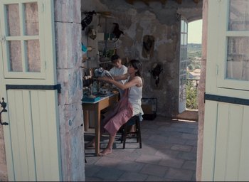 Movie still from “La Belle Noiseuse” (1991), directed by Jacques Rivette – A woman sitting at a table with a man standing next to her; Wide shot, High angle
