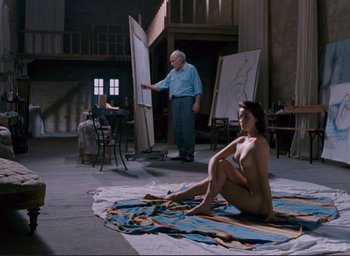 Movie still from “La Belle Noiseuse” (1991), directed by Jacques Rivette – A naked woman sitting on a rug in front of an easel; Wide shot, High angle